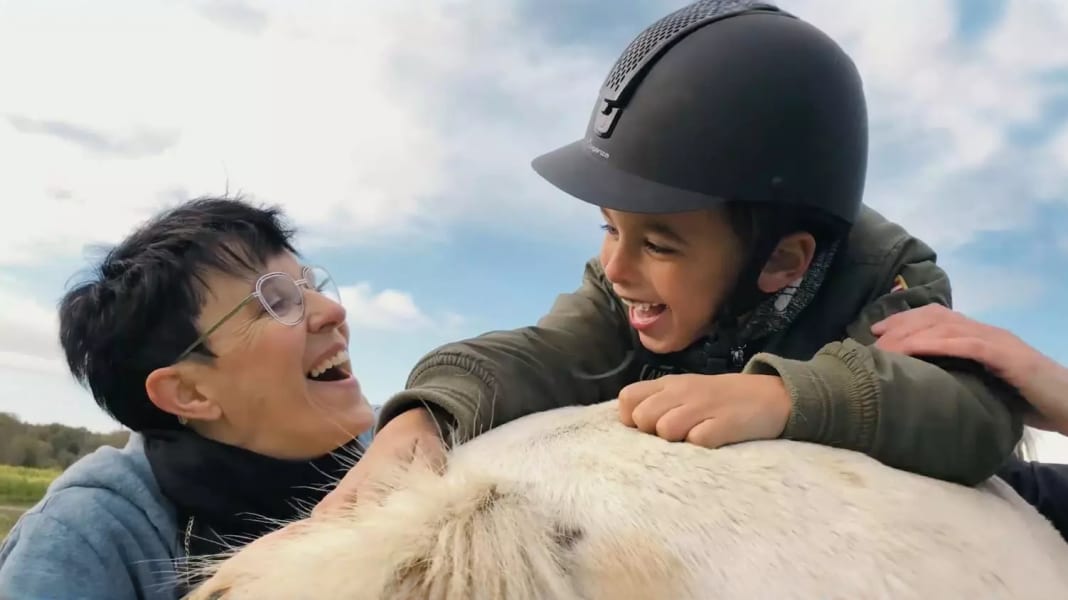 Jongetje met een helm op een paard met een lachende vrouw erbij.
